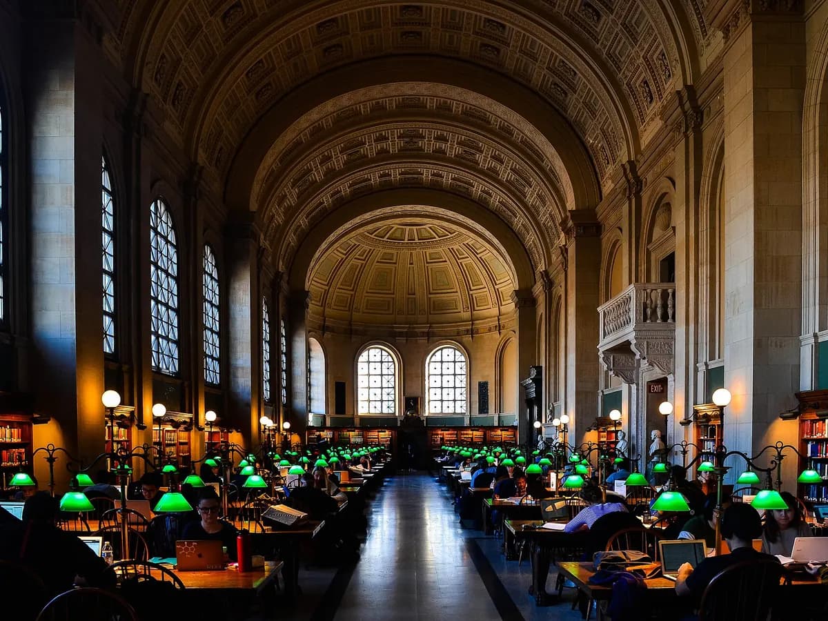 Bates Hall reading room with its coffered barrel vault, arched windows, long wooden tables and rows of glowing green lamps
