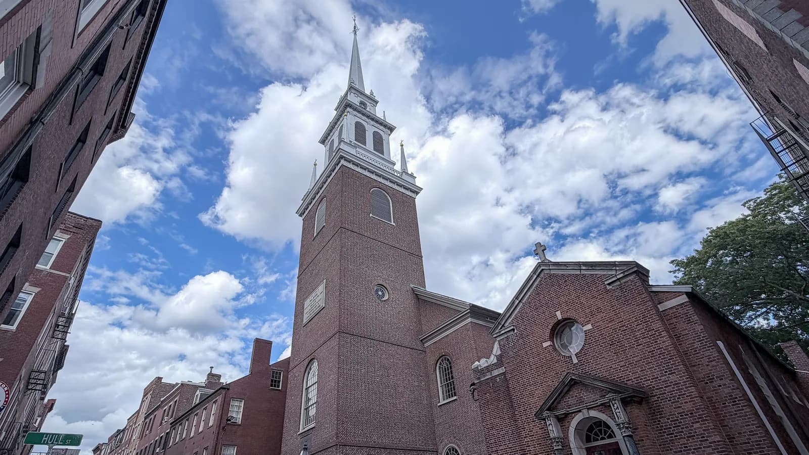 White steeple and red brick tower of the Old North Church rising above North End rowhouses under a blue sky with white clouds