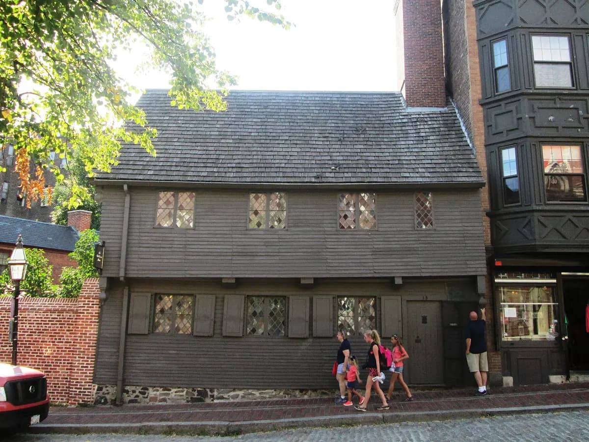 Dark gray clapboard Paul Revere House with diamond paned windows, shutters and an overhanging second story, as a family walks past on the brick sidewalk