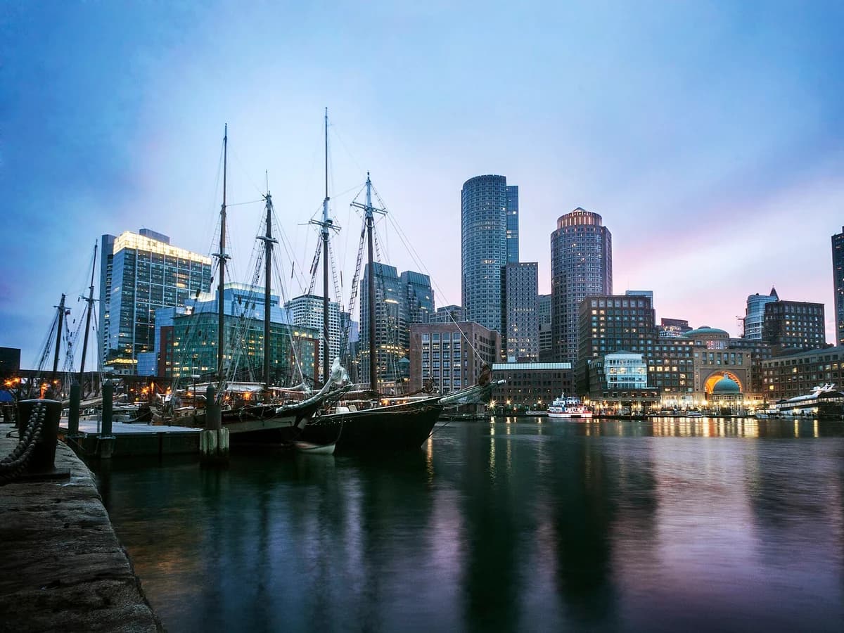 Tall masted ships moored at the Fan Pier docks at dusk, with the Financial District towers and the Rowes Wharf arch reflected in the harbor