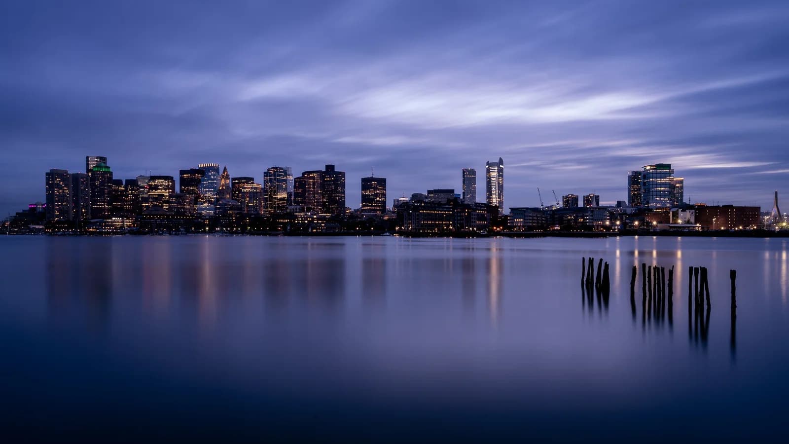 Downtown Boston skyline across a glassy long exposure harbor under purple clouds, seen from LoPresti Park in East Boston, with old pier pilings in the water