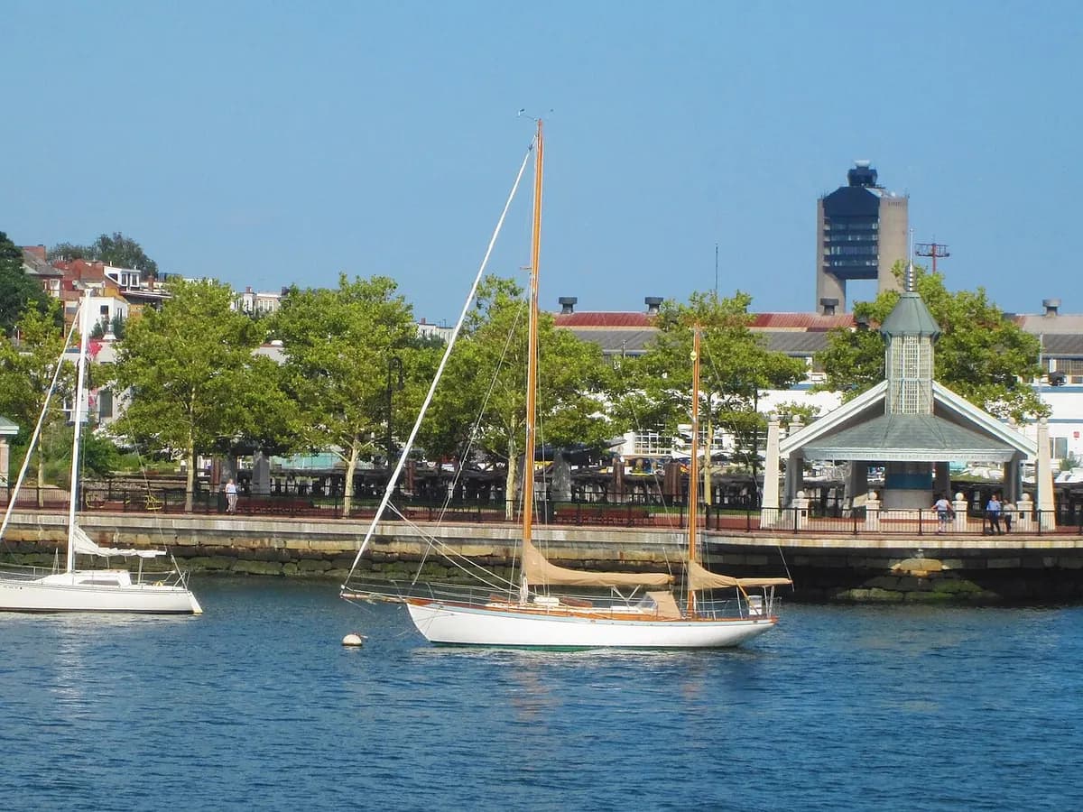 Sailboats moored off Piers Park, with its tree lined waterfront promenade, a green roofed pavilion and the Logan Airport control tower behind