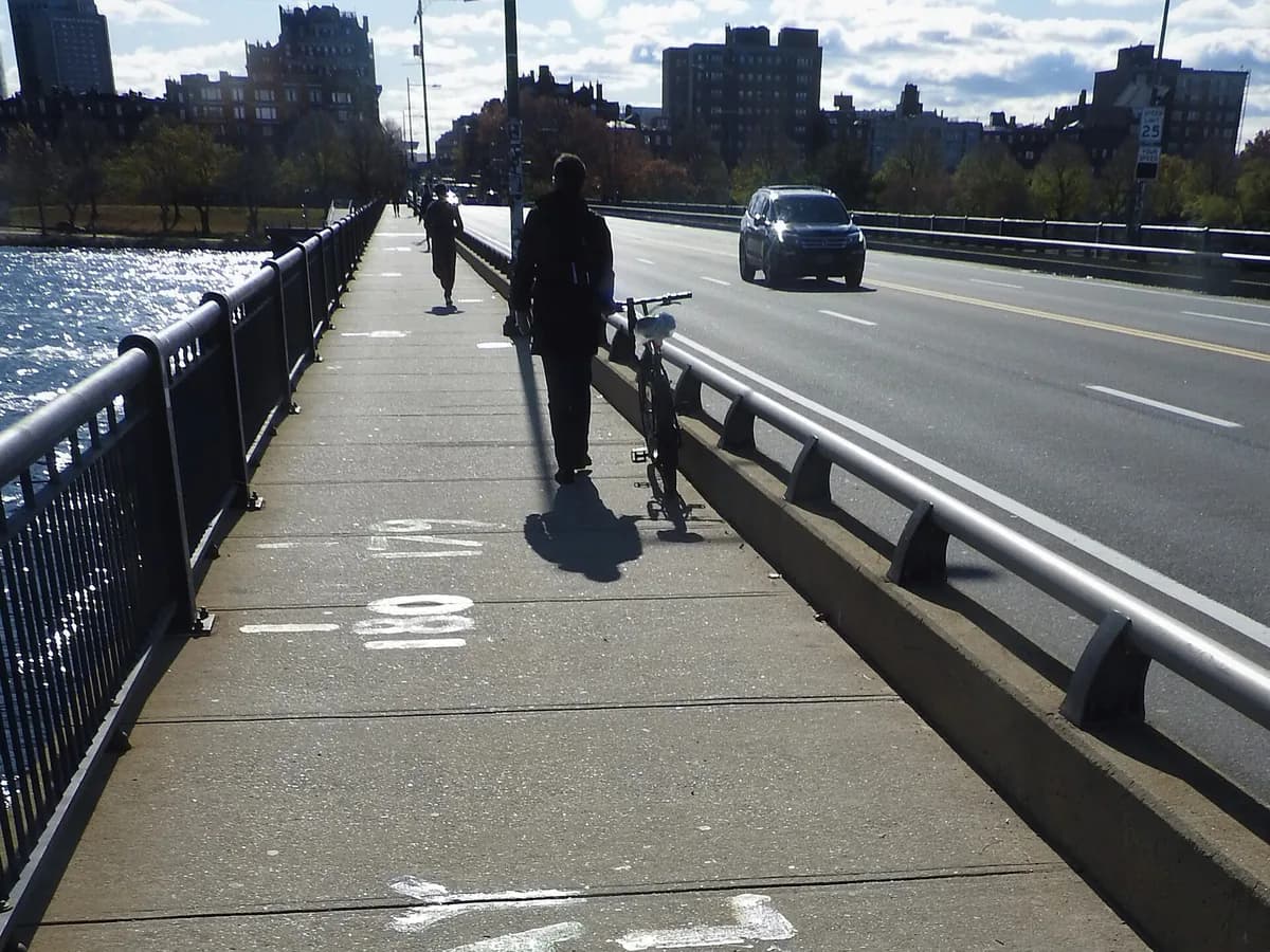 Sidewalk of the Harvard Bridge with painted Smoot length marks, a walker heading toward Back Bay apartment buildings and the sunlit Charles River beside the railing