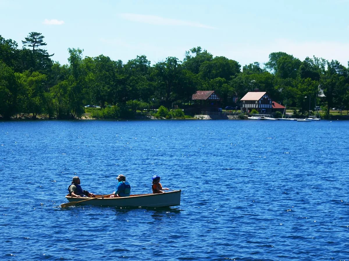 Three people rowing a small boat across blue Jamaica Pond, with a red roofed boathouse and dense green trees along the far shore