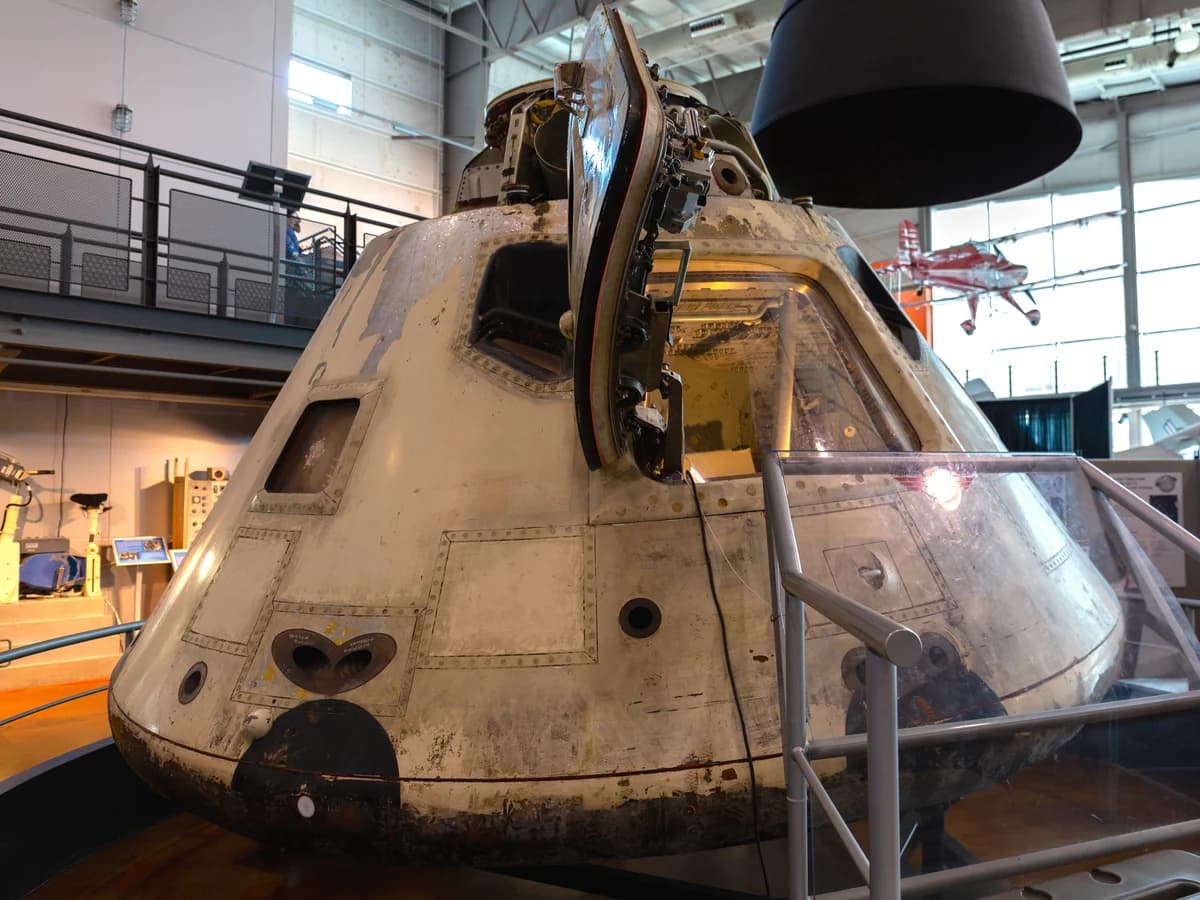 The weathered cone-shaped Apollo 7 command module, its hatch open, on display behind a metal railing and glass panel in a museum hangar