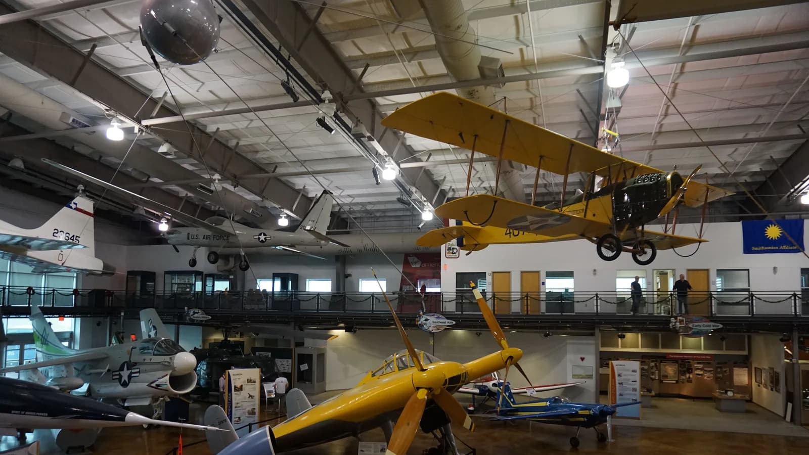 Vintage airplanes, including a yellow biplane, hang from the steel roof of a museum hangar above a yellow racing plane and other aircraft on a polished floor