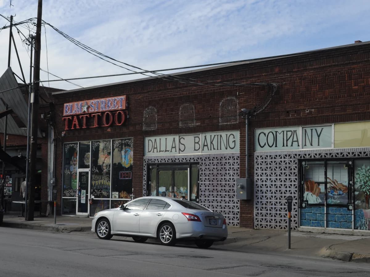 Low brick storefronts on Elm Street in Deep Ellum, with a neon Elm Street Tattoo sign and a Dallas Baking Company sign over patterned block screens