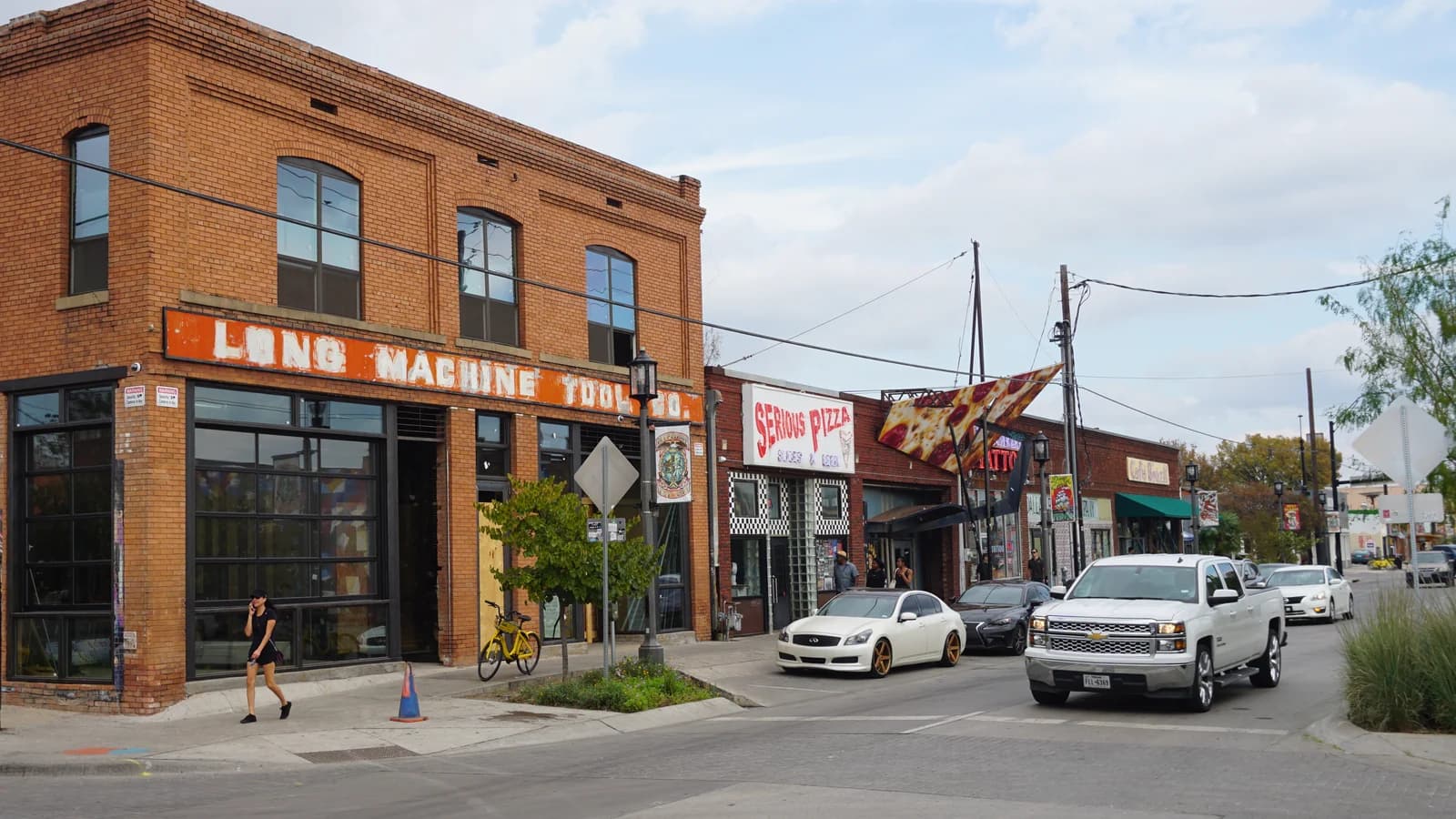 A red brick corner building with an old Long Machine Tool Co. sign on Elm Street in Deep Ellum, beside a Serious Pizza sign, parked cars and a pickup truck