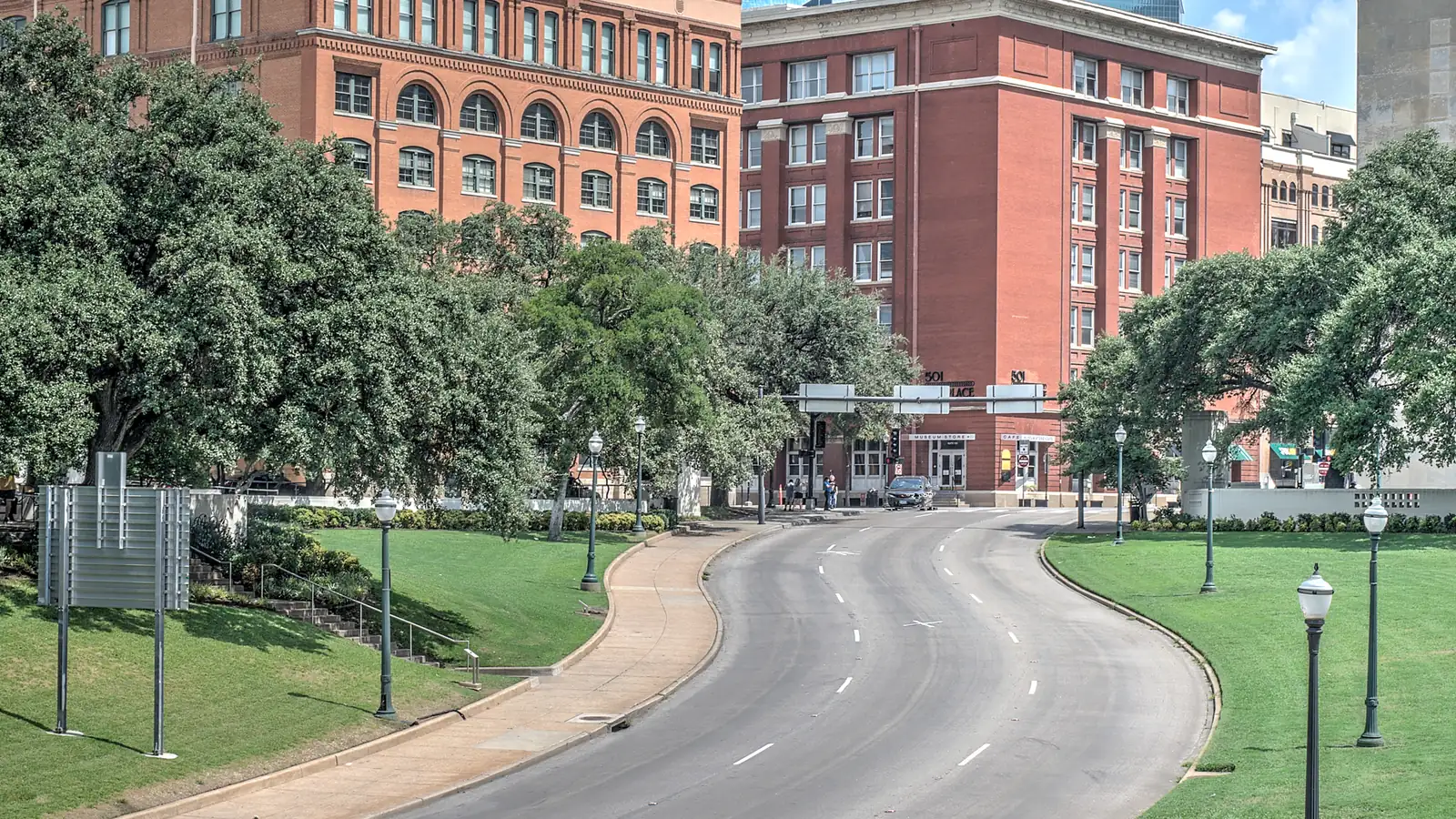 Elm Street curves downhill between green lawns and lamp posts toward the red brick former Texas School Book Depository and its neighbor, under a blue sky