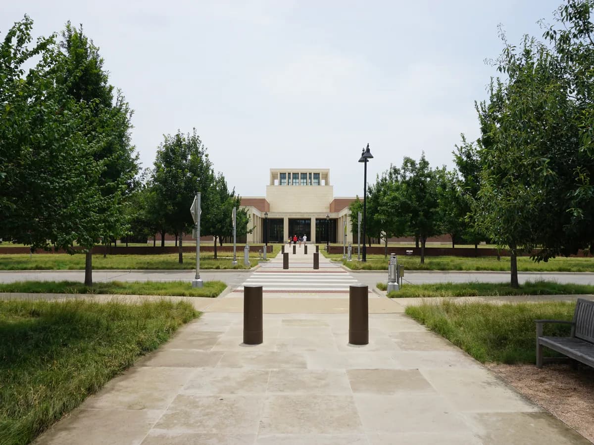 A wide stone walk with bollards and a bench leads through young trees and native grasses to the pale stone Bush Presidential Library and its lantern tower