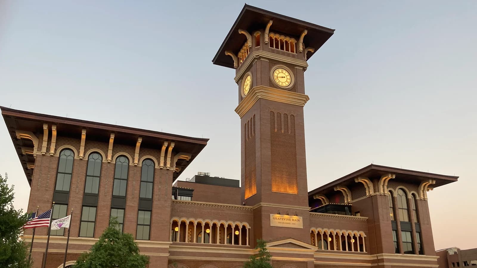 The brick clock tower of Grapevine Main at dusk, with a wide bracketed roof and lit clock faces, rising between two wings of tall arched windows