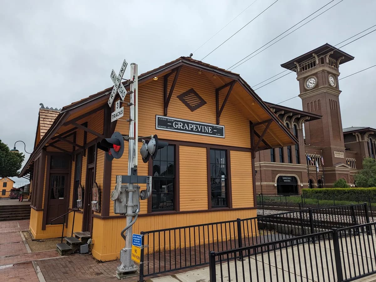 The yellow wooden Grapevine depot with a railroad crossing signal, and behind it the brick clock tower of Grapevine Main with a Harvest Hall sign over an arched door