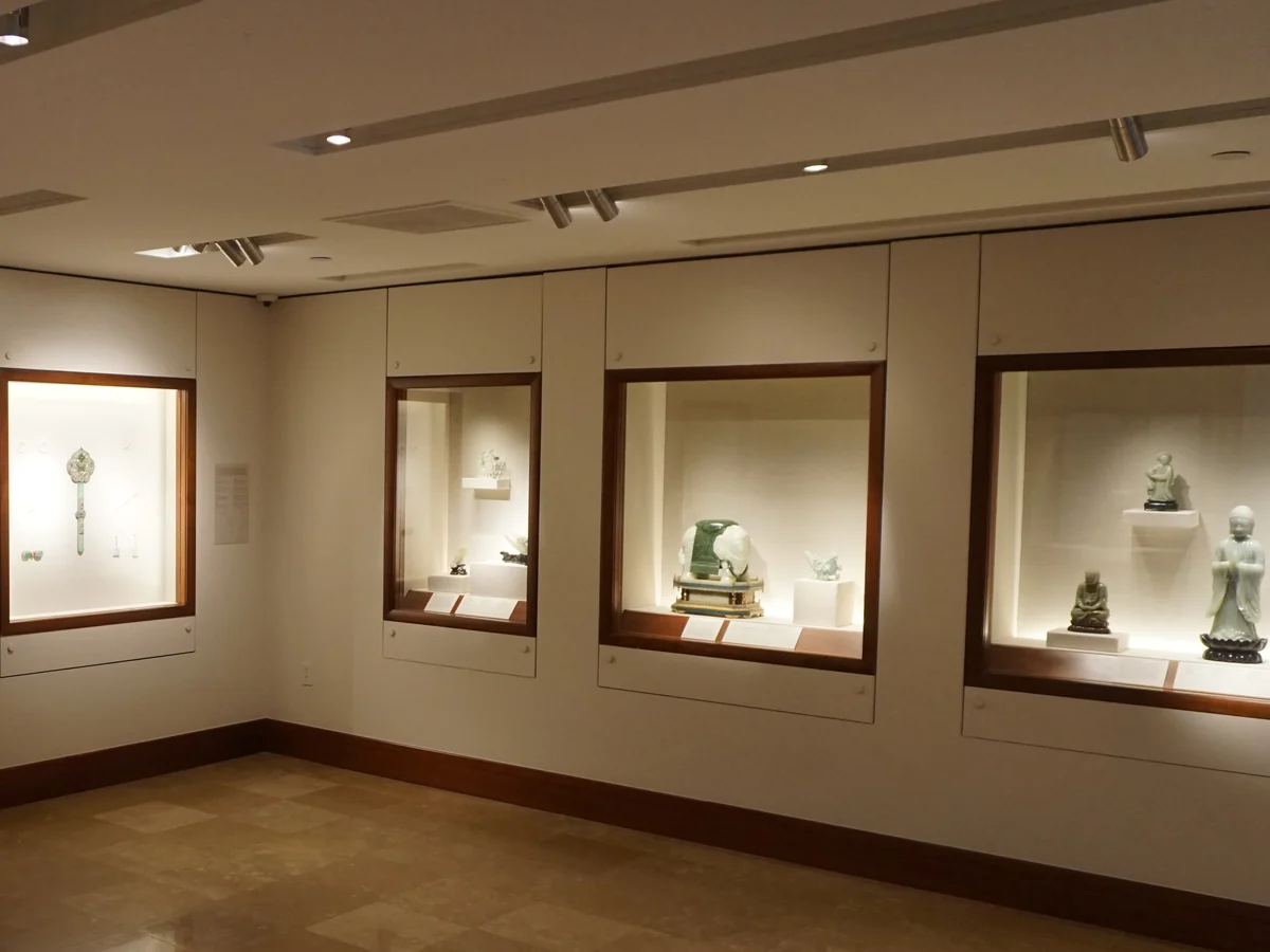 Lit wall cases in a quiet museum gallery display carved jade pieces, including a green and white jade carving on a stand and pale green seated and standing figures