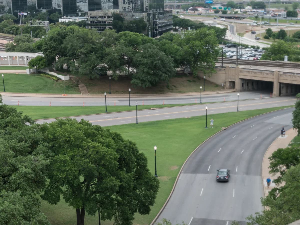 Elm Street curves past a wide lawn and lamp posts toward the triple underpass, seen from high up in the old book depository, with trees and Dallas towers beyond