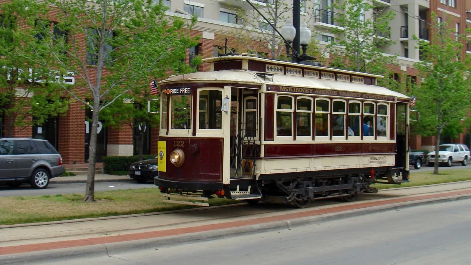 A maroon and cream vintage M-Line streetcar, car 122, on a grassy median track beside young trees and brick apartment buildings in Uptown Dallas