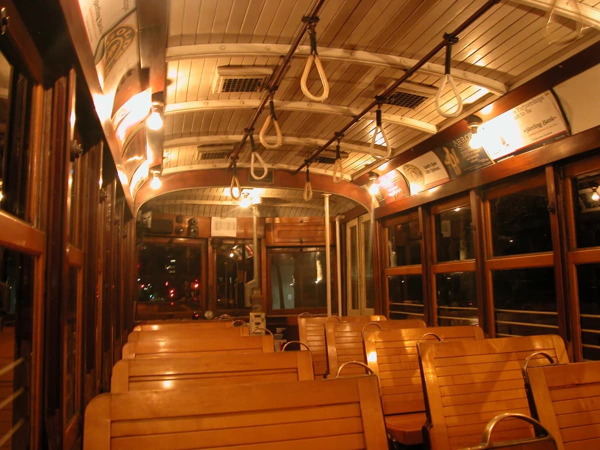 Inside a vintage streetcar at night, with rows of polished wooden bench seats, hanging hand straps and warm lights under a curved wooden ceiling