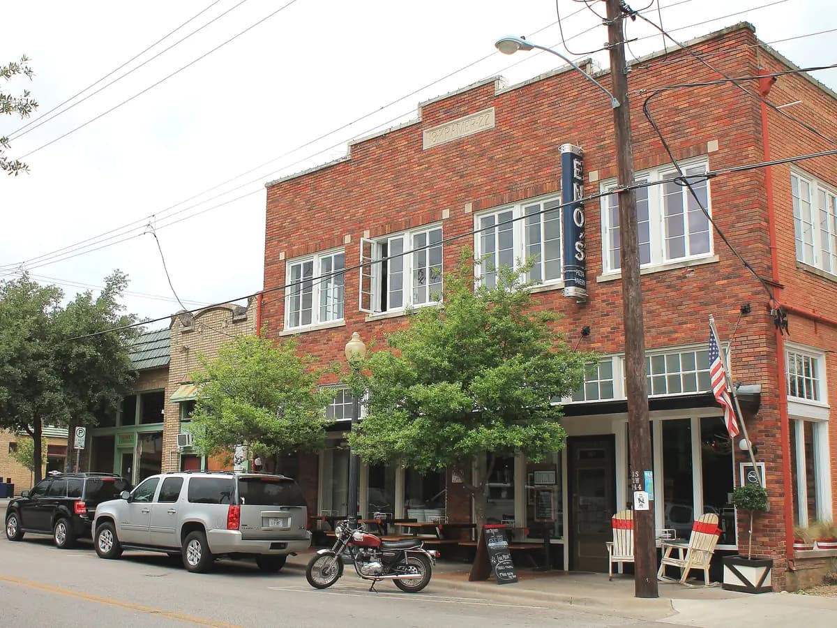 A two story red brick building with tall white windows on Bishop Avenue, with leafy trees, sidewalk tables, porch chairs and a motorcycle parked out front