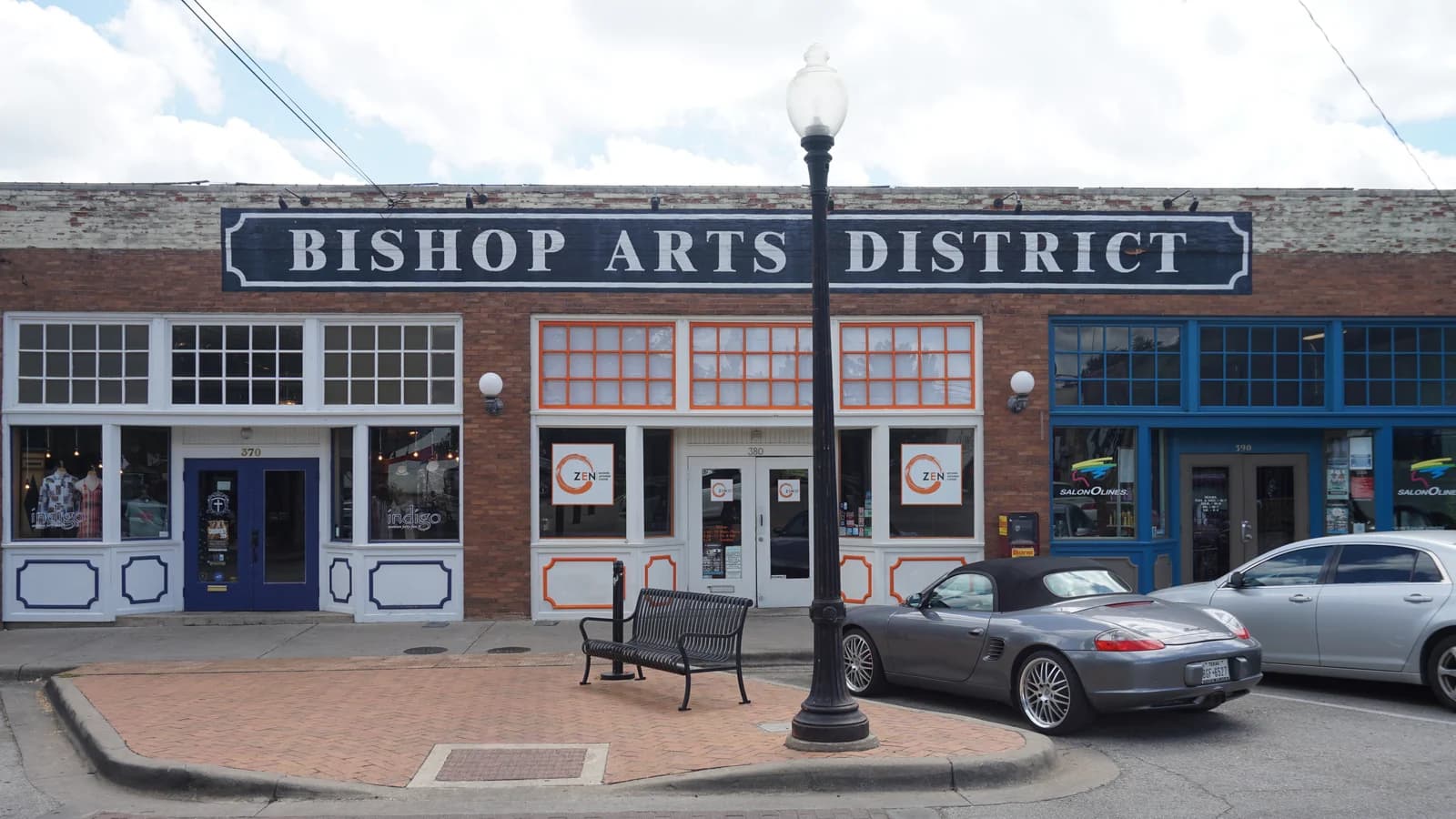 A long brick storefront under a big Bishop Arts District sign, with a bench and street lamp on a brick corner and a silver convertible parked out front
