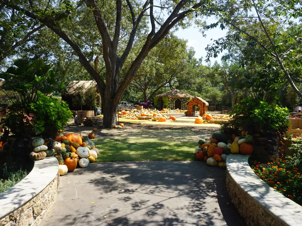 Pumpkins and gourds piled along a stone-edged garden path, leading to pumpkin-covered cottages and an arch under tall shade trees