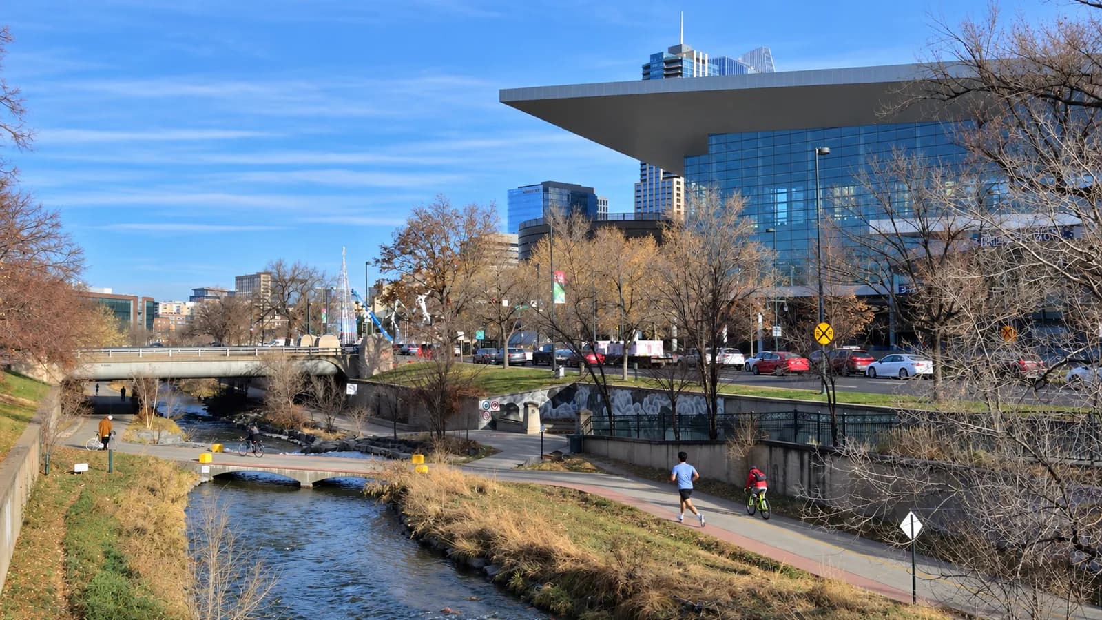 A man in a light blue tee runs on the Cherry Creek Trail toward low bridges, beside the creek, bare trees and a cantilevered glass building with towers behind