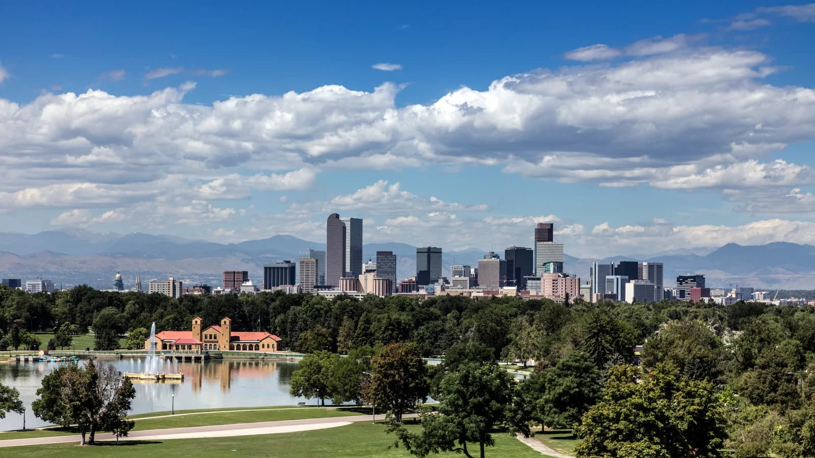 The Denver skyline beyond City Park's trees and Ferril Lake, with the red-roofed boathouse, a fountain and the Front Range under white clouds