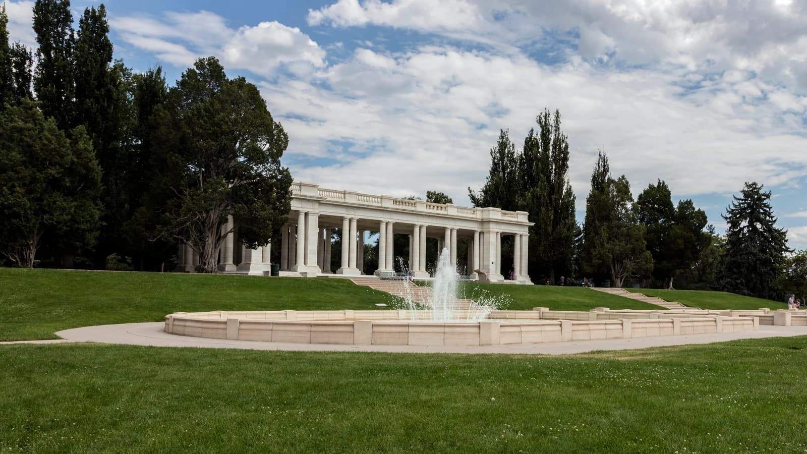 The white marble Cheesman Memorial Pavilion with its columns, above a lawn and a fountain pool, framed by tall dark evergreens under a cloudy sky