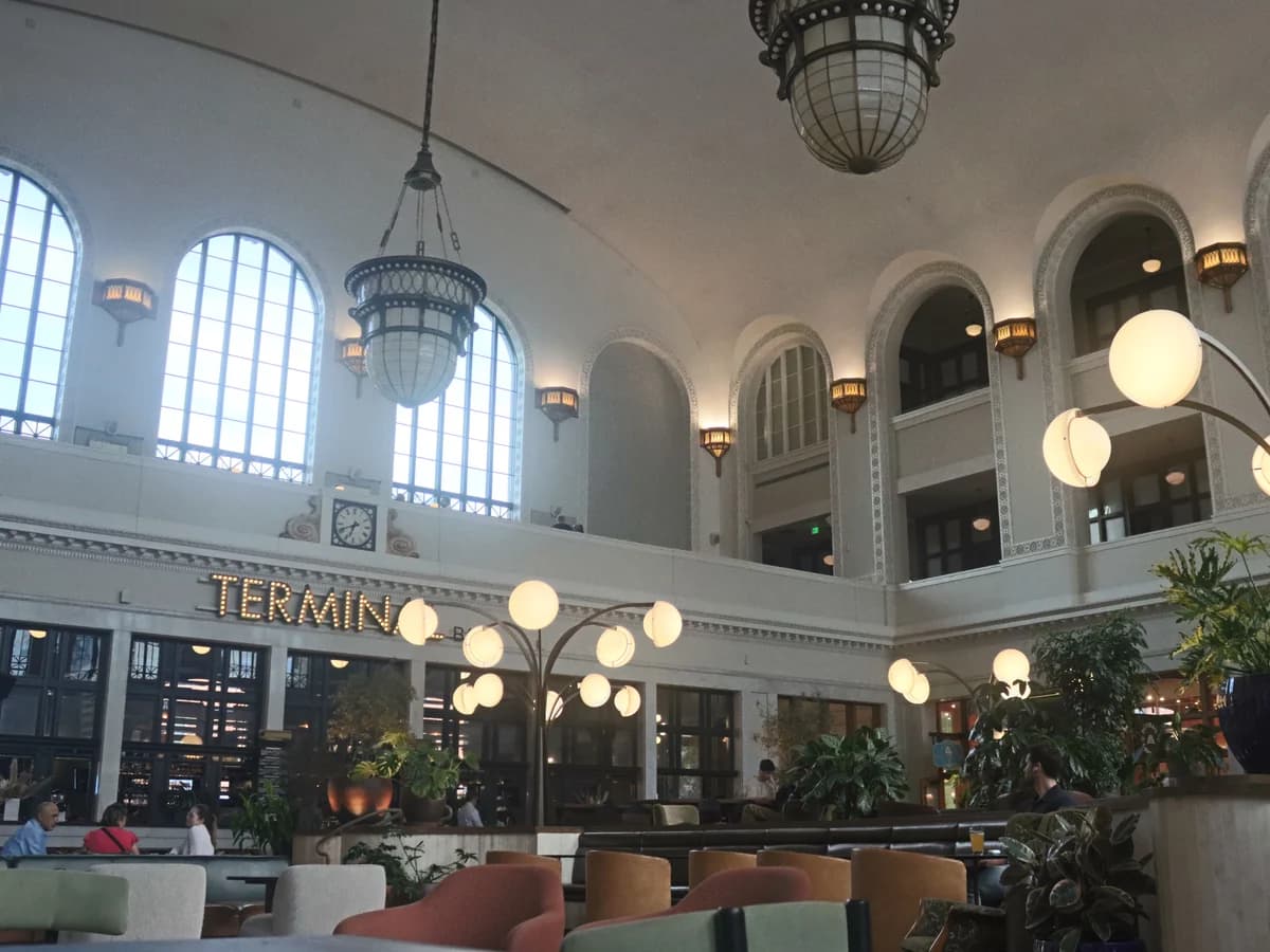 The Great Hall of Denver Union Station, with tall arched windows, hanging lanterns, globe lamps and lounge chairs below the lit Terminal Bar sign