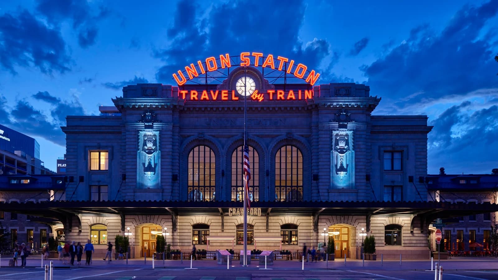 Denver Union Station's granite front at dusk, its red neon Union Station and Travel by Train sign glowing above three tall arched windows