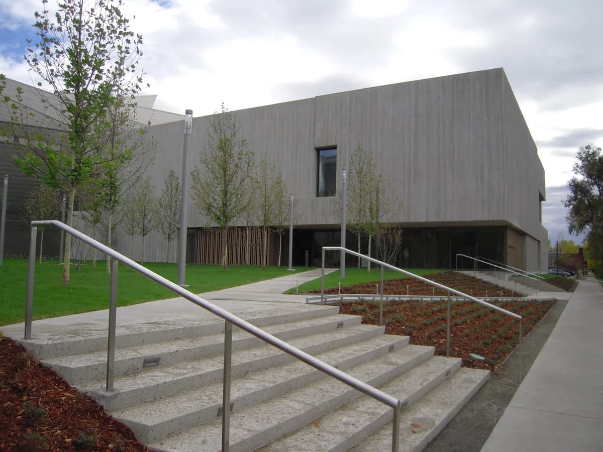 The gray concrete Clyfford Still Museum behind young trees and a lawn, with wide stone steps and metal handrails in the foreground under a cloudy sky