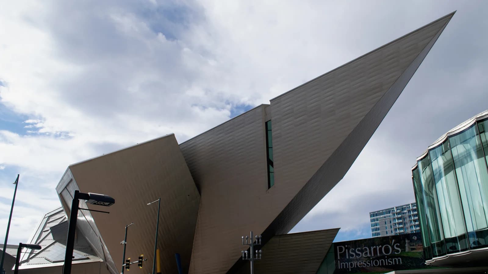 The Hamilton Building's sharp, angled titanium-clad forms jut out over a plaza beside a curved glass wall, under a partly cloudy sky