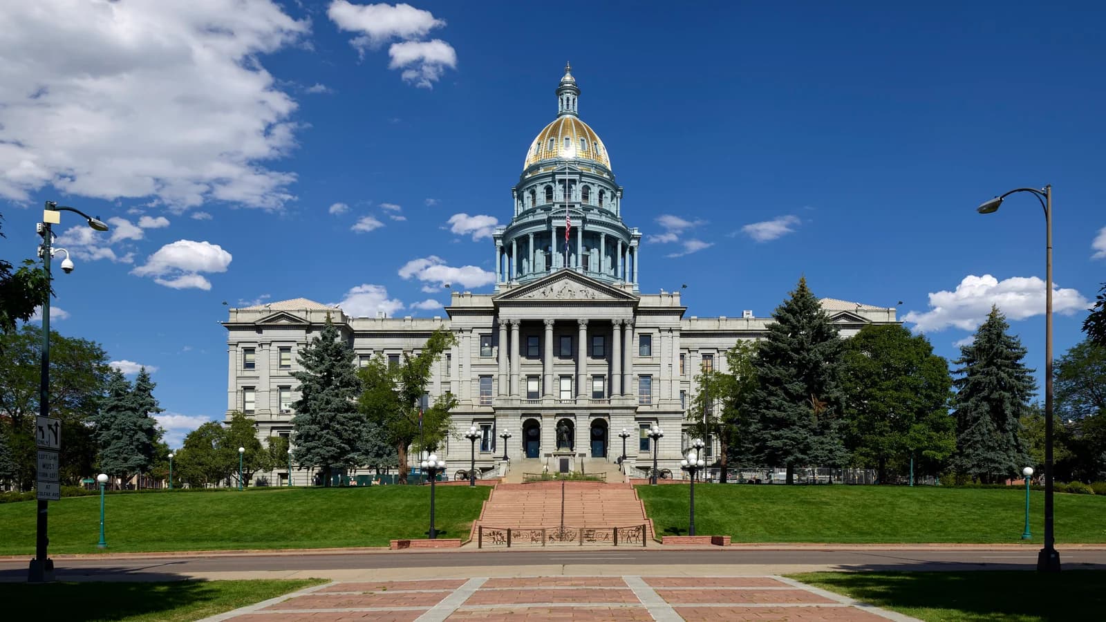 A brick-paved walk crosses Civic Center Park's lawns toward the gold-domed Colorado State Capitol, framed by evergreens and lamp posts under a blue sky
