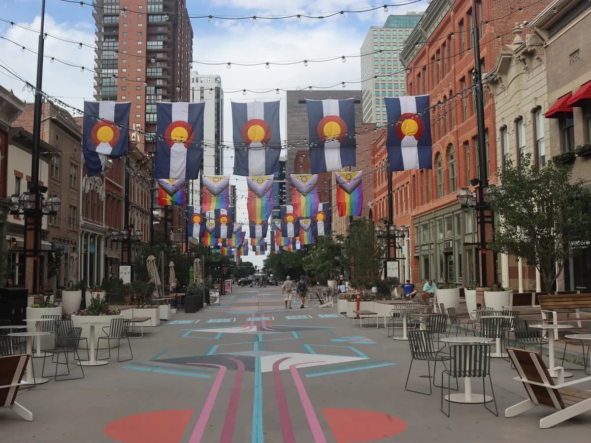 Larimer Square's pedestrian street under strings of lights and rows of Colorado flags, with brick Victorian buildings, cafe chairs and a painted pavement