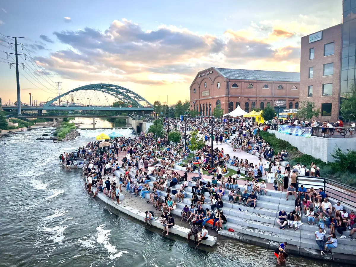 Crowds sit on Confluence Park's stone terraced steps above the South Platte's rapids at sunset, beside a brick building and an arched bridge