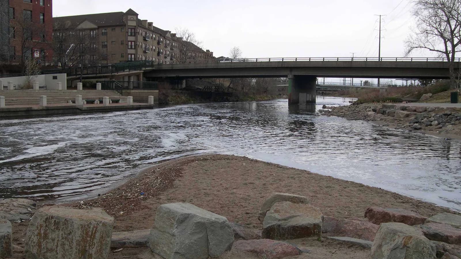 Cherry Creek pours into the South Platte River over a rocky bank at Confluence Park, with a road bridge, brick buildings and bare trees behind