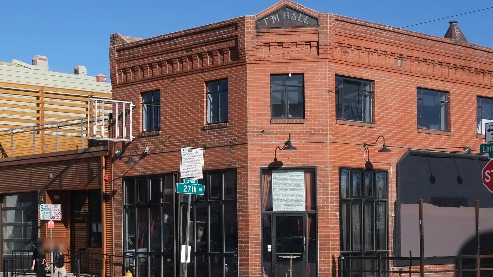 The red brick F.M. Hall building on the corner of Larimer and 27th Street in RiNo, with tall dark storefront windows and wall lamps under a blue sky