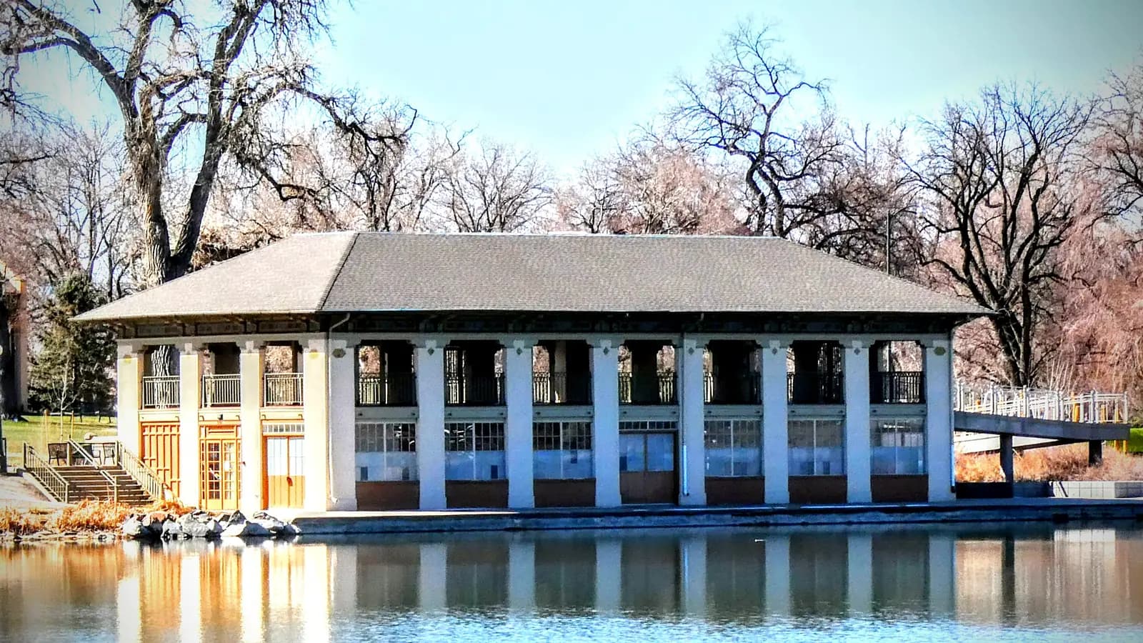 The pale columned Washington Park boathouse on the shore of Smith Lake, reflected in calm blue water, with bare spring trees behind it