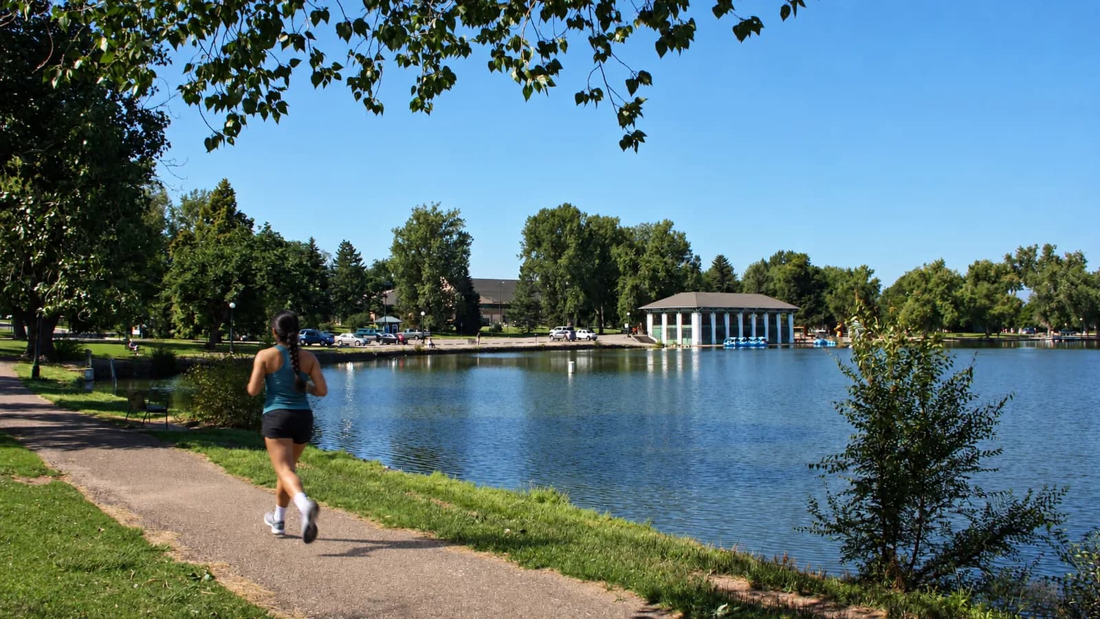 A woman with a long braid runs on a paved path along Smith Lake, with leafy trees and a white boathouse across the water under a clear blue sky