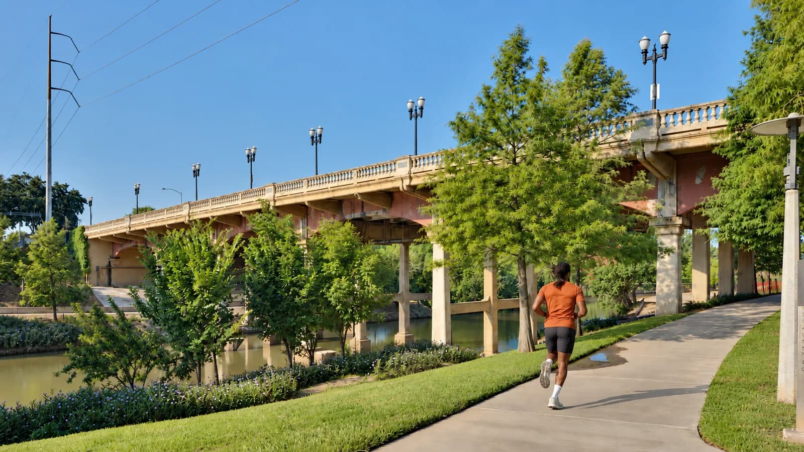 A runner in an orange tee follows a curving paved path along Buffalo Bayou beside the old Sabine Street Bridge, with lamp posts and young trees under a blue sky
