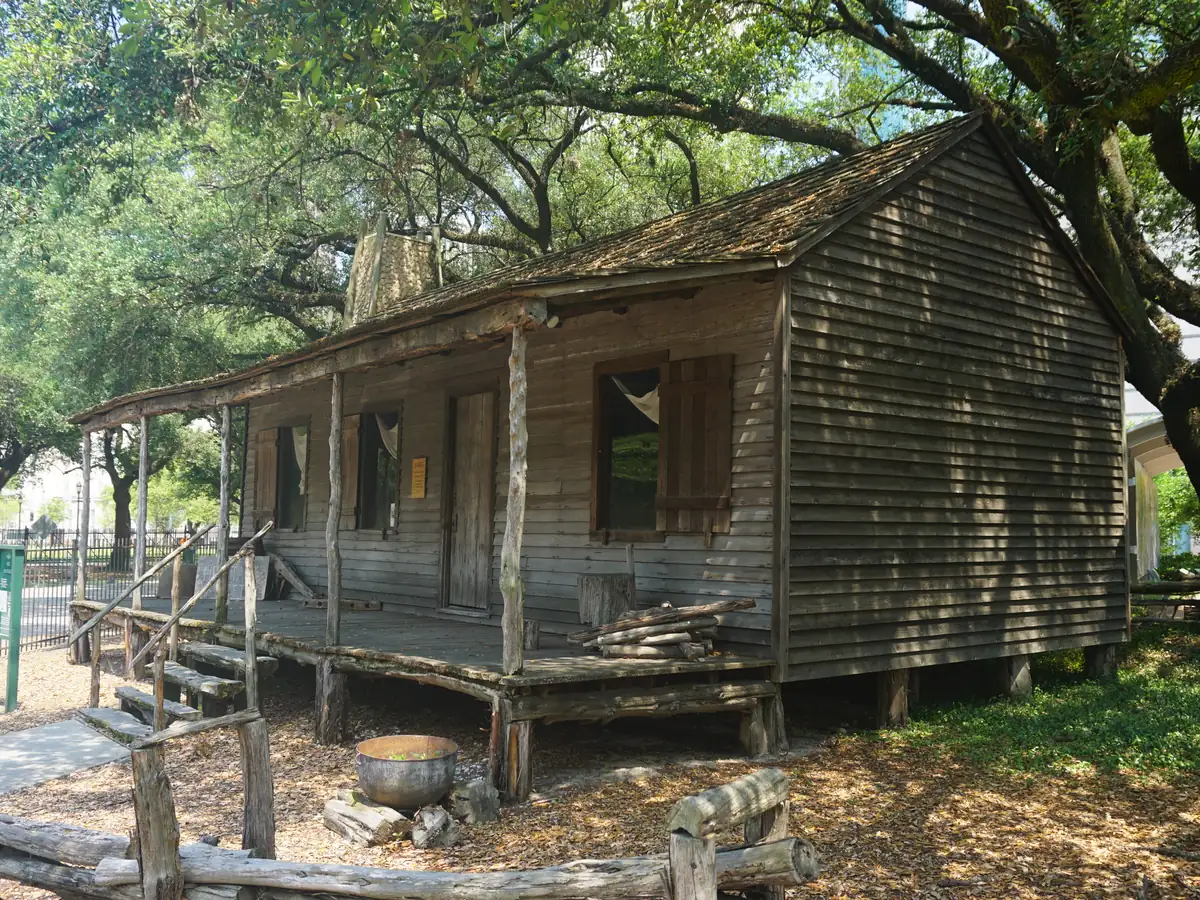 A weathered wooden log cabin with a shingled porch roof and a stone hearth beside it, shaded by large trees on a sunny day