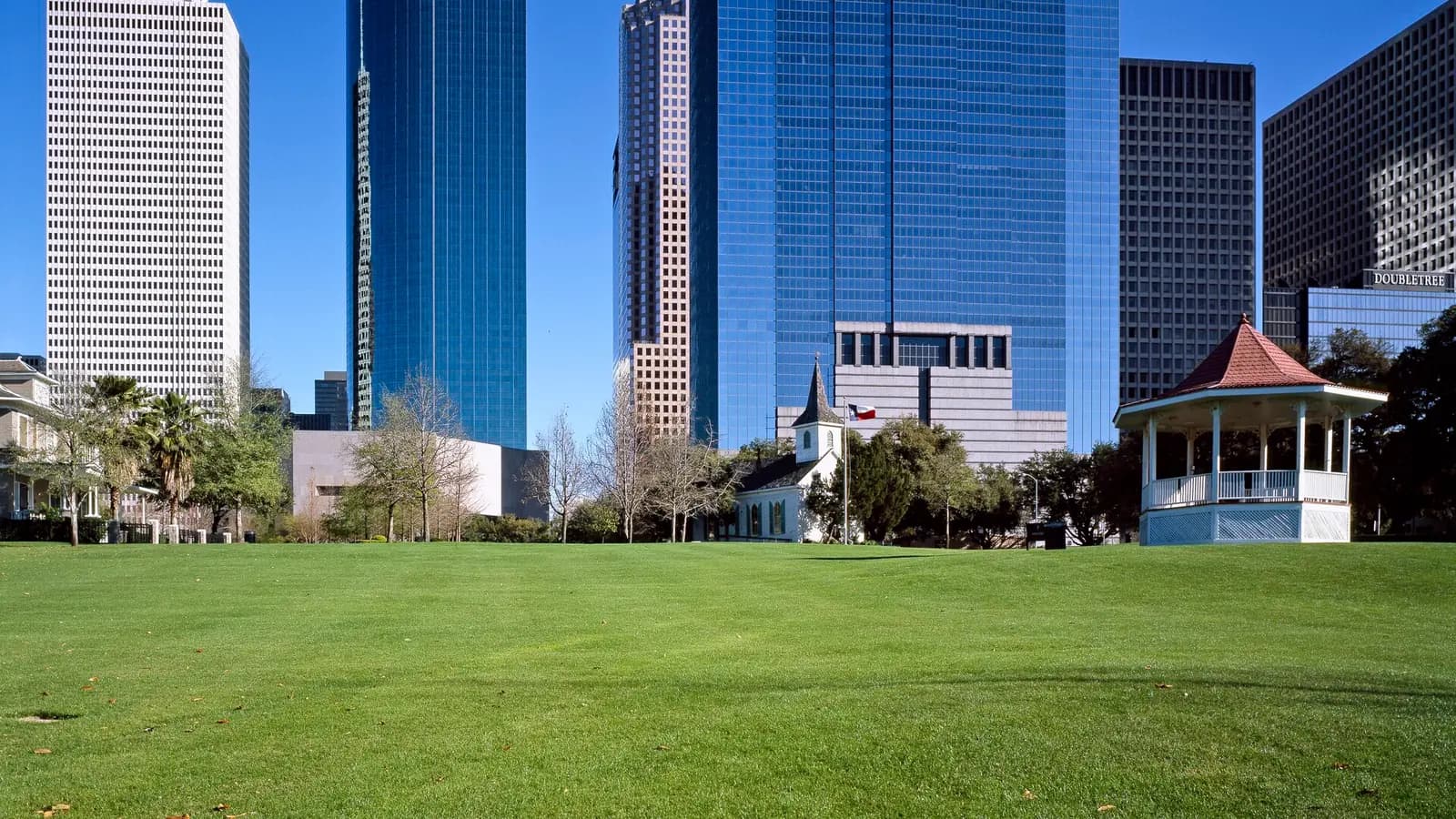 A wide green lawn with a white gazebo and a small white wooden church, with tall glass office towers rising behind the trees under a clear blue sky