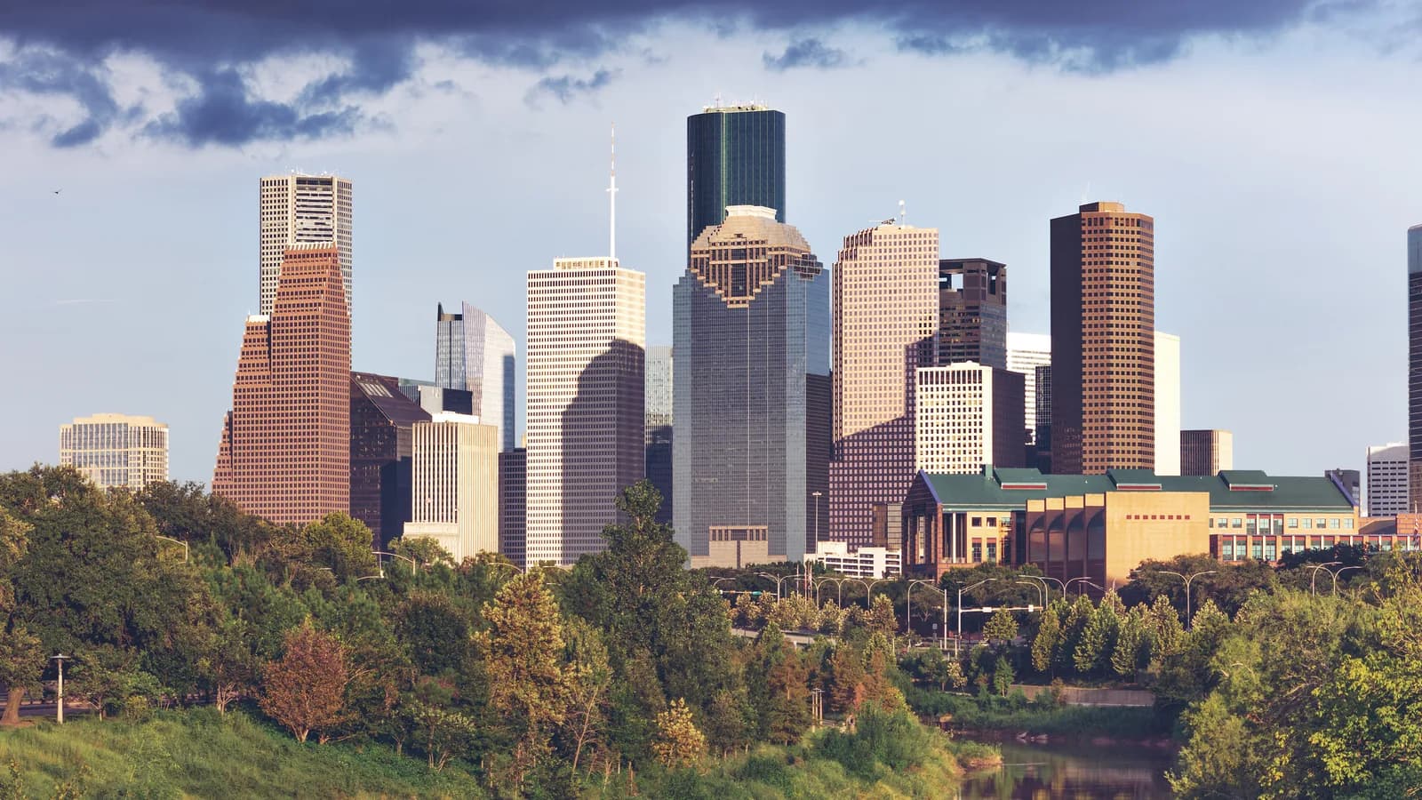 Downtown Houston's towers lit by low sun under dark storm clouds, rising above the green treetops of Buffalo Bayou Park and the bayou