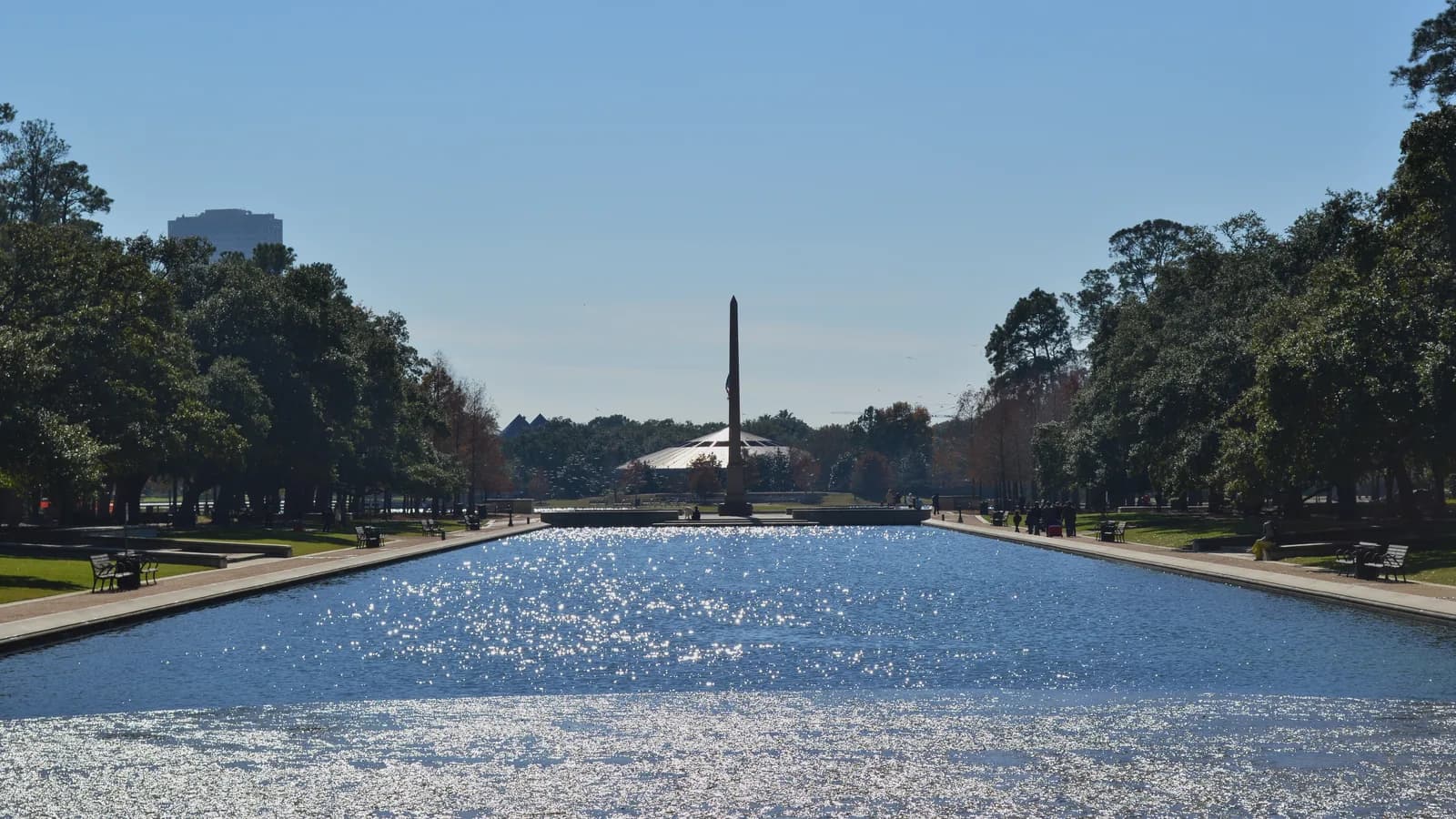 A long sparkling reflecting pool between rows of live oaks and park benches, leading to a tall obelisk and a domed pavilion under a clear sky