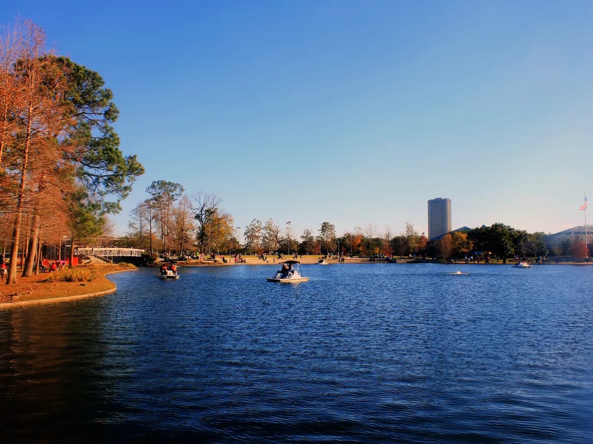 A calm blue park lake with pedal boats out on the water, autumn trees on the shore, a small footbridge and a lone tower in the distance