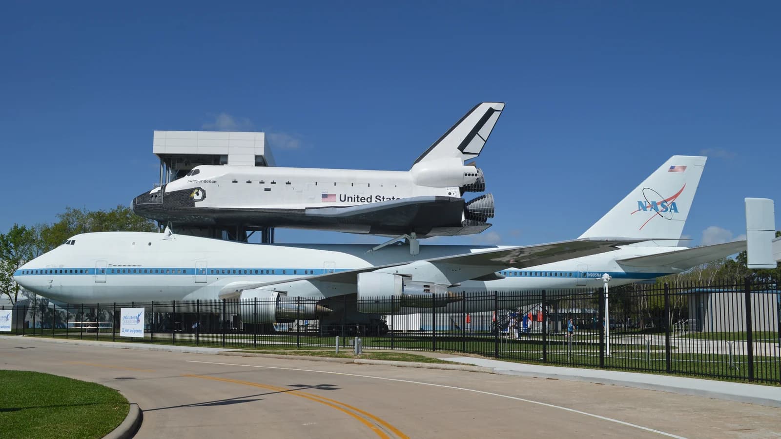 A white space shuttle replica mounted on top of a NASA Boeing 747 on tall supports behind a fence, with a tower and trees under a deep blue sky