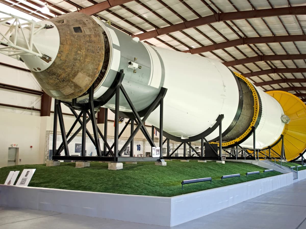 A huge white Saturn V rocket lying on its side on black supports inside a long hall, with the capsule end in front and its stages stretching away