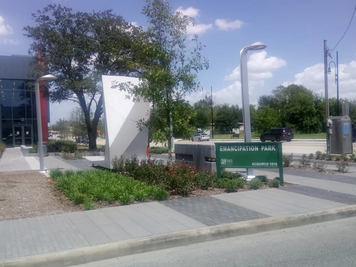 The entrance to Emancipation Park, with a green park sign, a tall white angled marker, young trees and flower beds beside a modern glass building