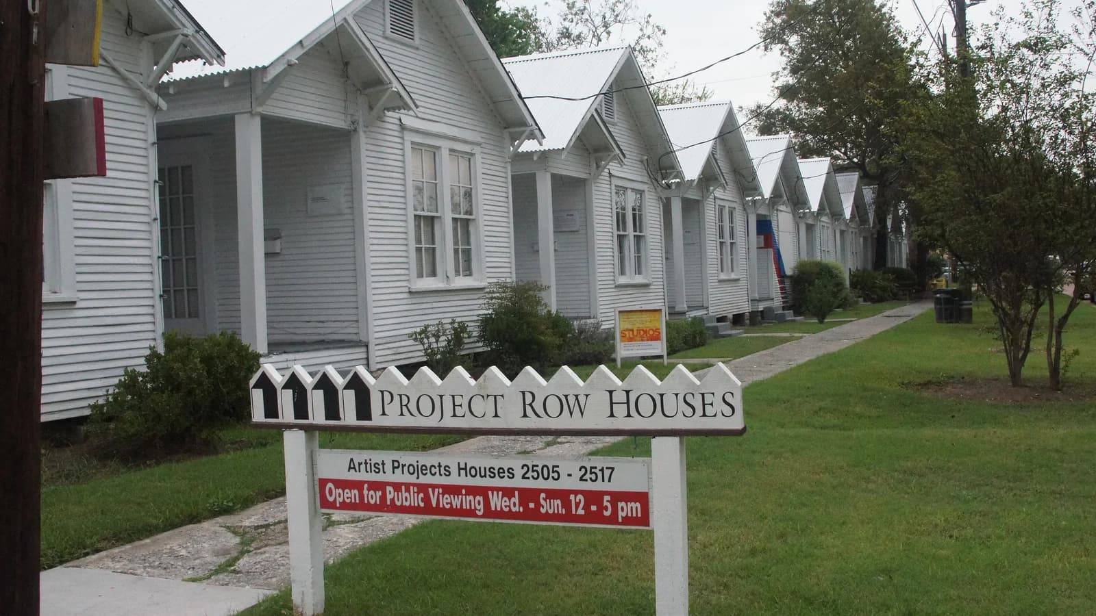 A row of white wooden shotgun houses with front porches along a green lawn, with a Project Row Houses sign in front under a hazy sky