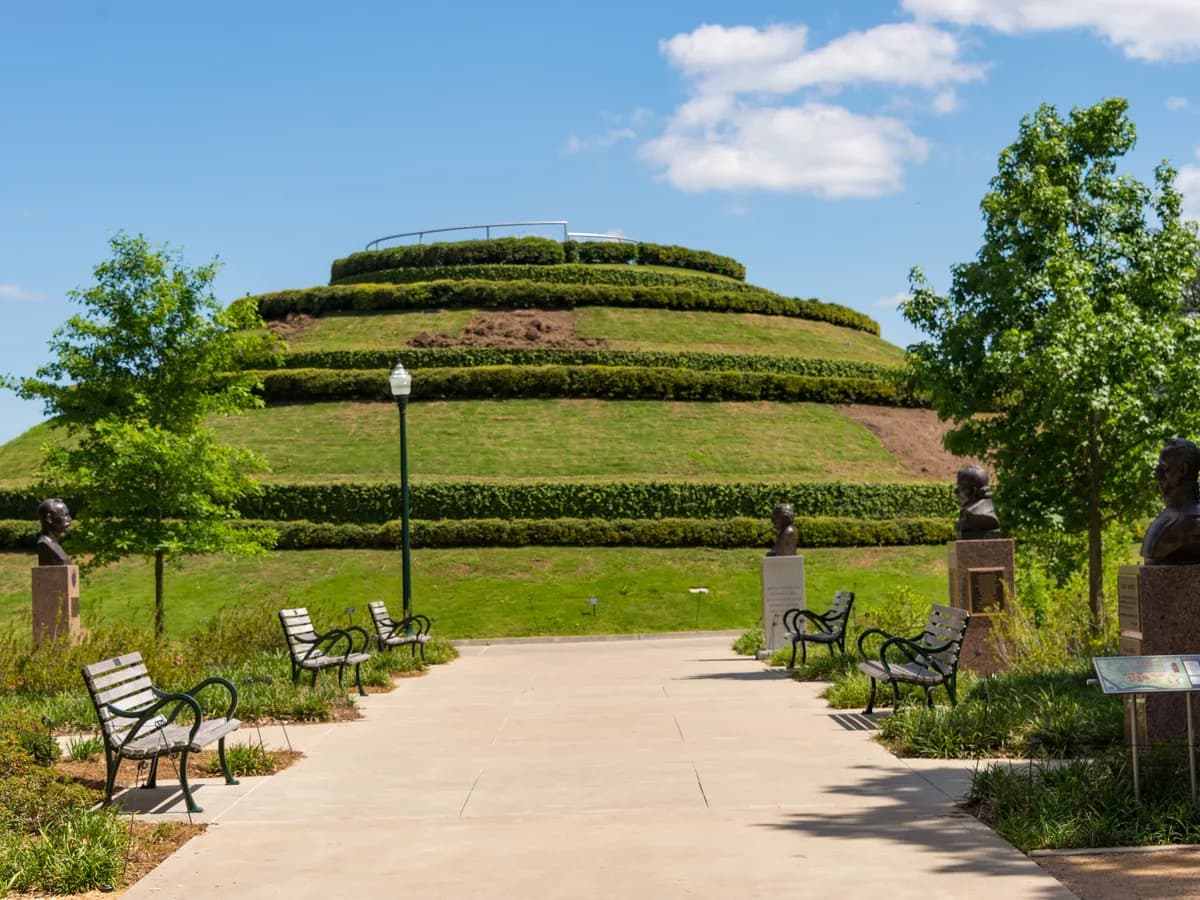 A terraced grassy mound with hedges ringing its sides, seen down a paved path lined with benches, young trees and small bronze busts on pedestals