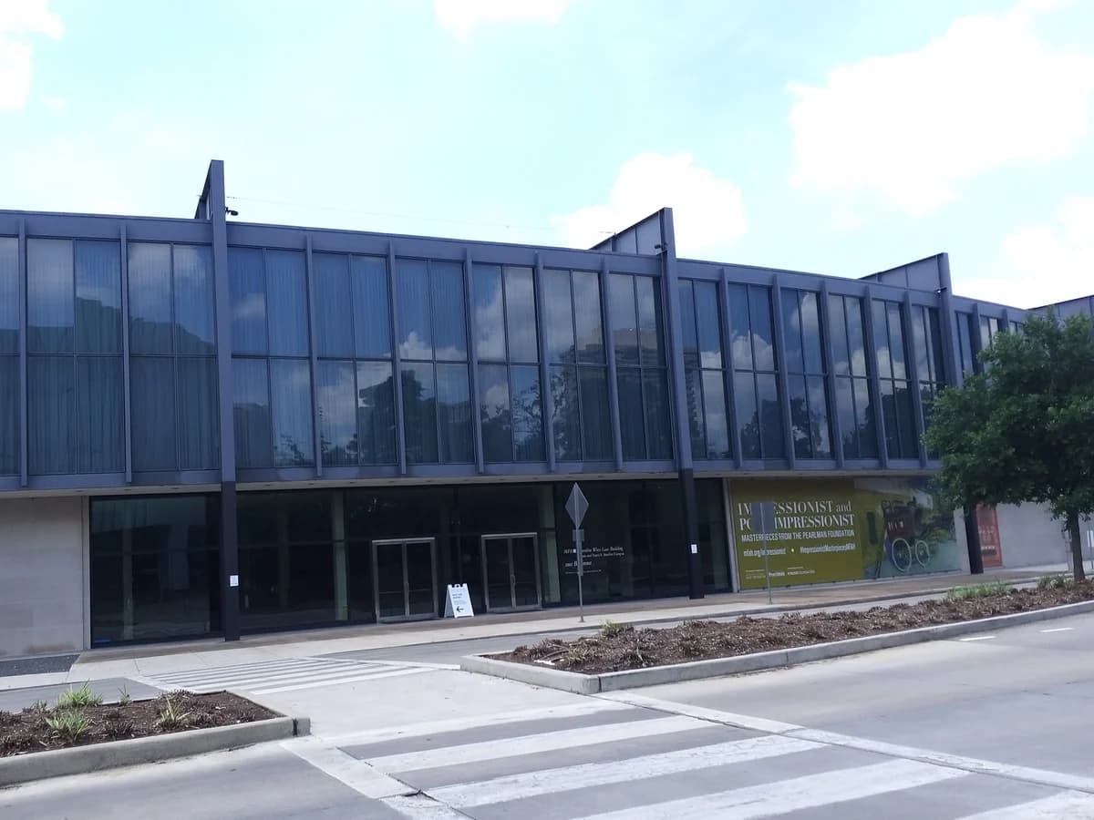 A long two story glass and steel museum building with a black frame, a crosswalk and planted median in front under a blue sky with light clouds