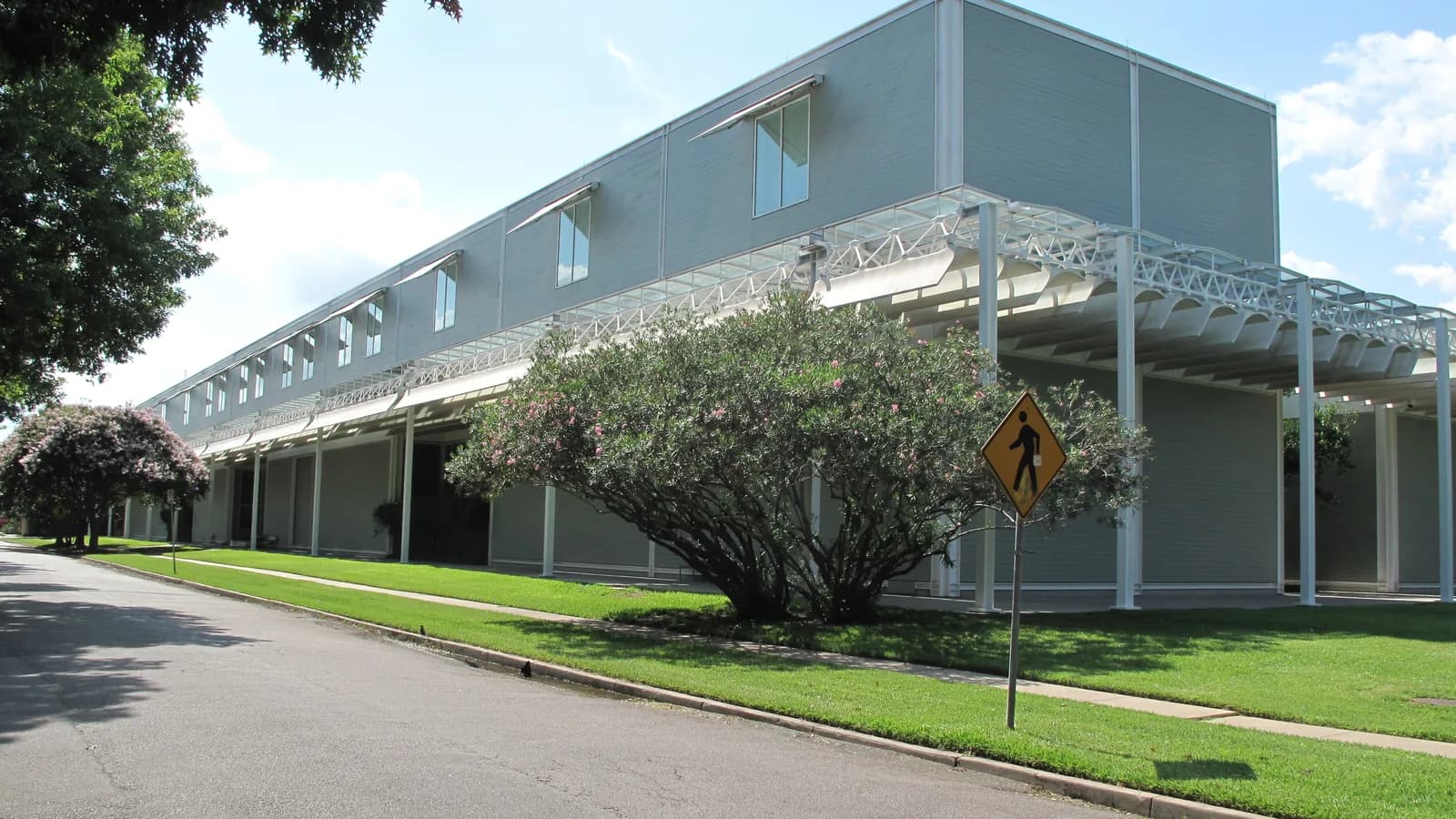 A long low museum building with gray clapboard walls and a white steel canopy along a lawn and sidewalk, with flowering shrubs and a quiet street